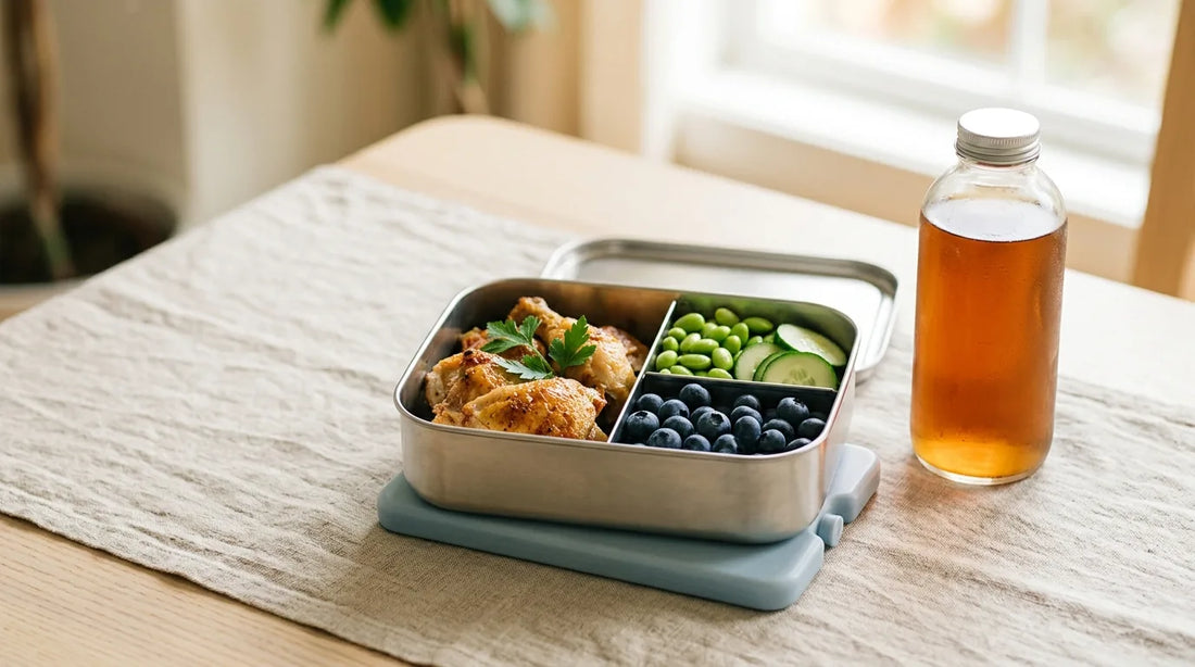 Open 3-compartment stainless bento box with roasted chicken, edamame, and blueberries beside a clear 18 oz glass bottle on a linen tablecloth