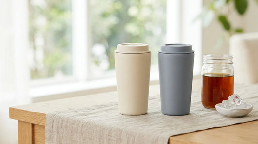 Two matte stainless steel vacuum-insulated travel mugs beside a glass jar of cold brew coffee and ice cubes on a linen-covered light wood table