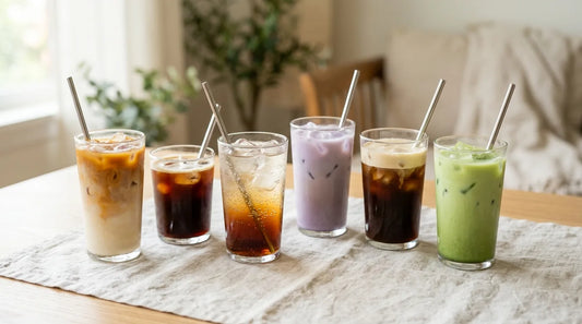 Six café-style iced drinks including iced latte, cold brew, sparkling tonic, and matcha latte arranged on a linen tablecloth over a light wood table
