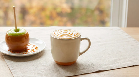 Caramel apple latte in a cream stoneware mug beside a glossy caramel-dipped apple with seasonal sprinkles on a light wood table with linen cloth