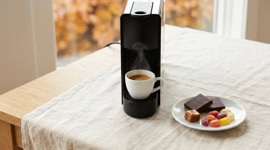 Matte-black espresso machine with a pulled espresso shot in a white ceramic cup beside dark chocolate squares and colorful candy pieces on a light wood table