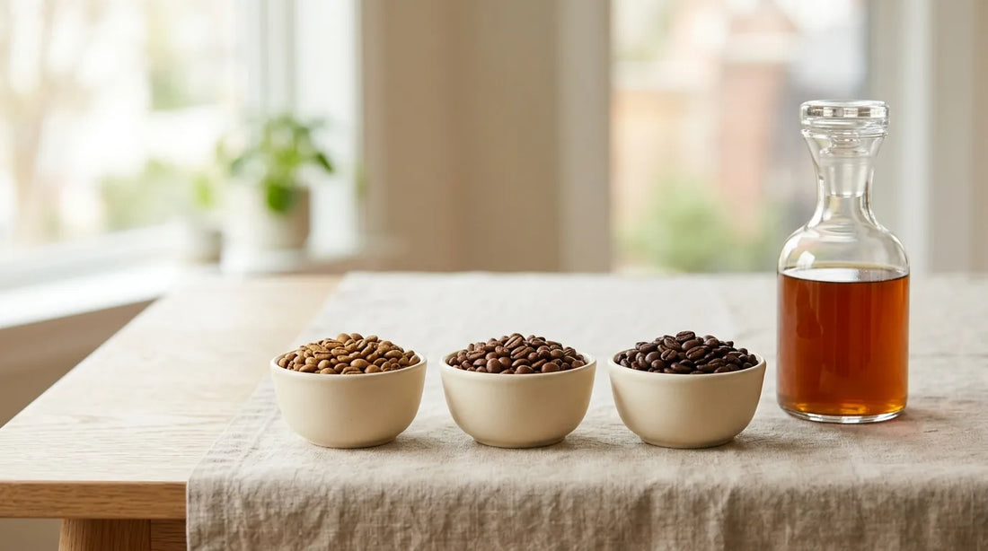 Three ceramic bowls of light, medium, and dark roast cold brew coffee beans beside a glass carafe of cold brew on a linen tablecloth