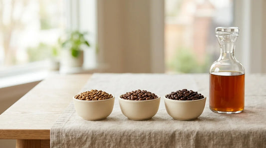 Three ceramic bowls of light, medium, and dark roast cold brew coffee beans beside a glass carafe of cold brew on a linen tablecloth