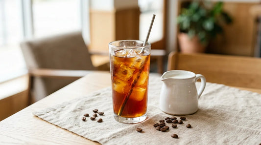 Tall glass of cold brew coffee over clear ice on a light wood table with linen cloth and a small white ceramic pitcher beside it