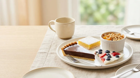 Four café-style easy summer desserts on a linen tablecloth — chocolate ganache tart slice, lemon bar, frozen yogurt bark, and peach crumble ramekin