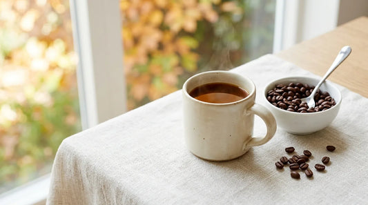 Warm cream stoneware mug of freshly brewed coffee beside a white ceramic bowl of whole roasted beans on a linen-covered light wood table for fall mornings