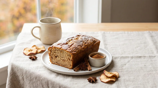 Golden-brown apple cinnamon loaf on a white ceramic plate with cinnamon sticks, star anise, and a warm cream ceramic mug on a linen tablecloth