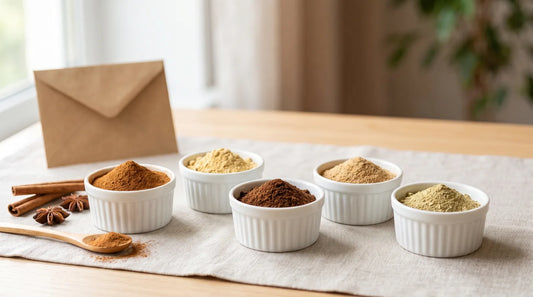 Five ceramic ramekins filled with fall baking spices — cinnamon, ginger, clove, nutmeg, and cardamom — on a linen tablecloth with cinnamon sticks and a wooden spoon