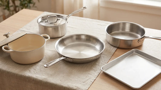 Five essential cookware pans arranged on a linen-covered light wood table: stainless skillet, saucepan with lid, sauté pan, cream Dutch oven, and half-sheet pan