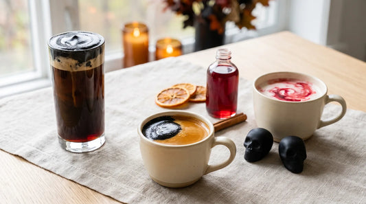 Halloween home café drink station featuring black foam espresso, blood-orange latte with crimson swirl, and dark cold brew on a linen-covered light wood table