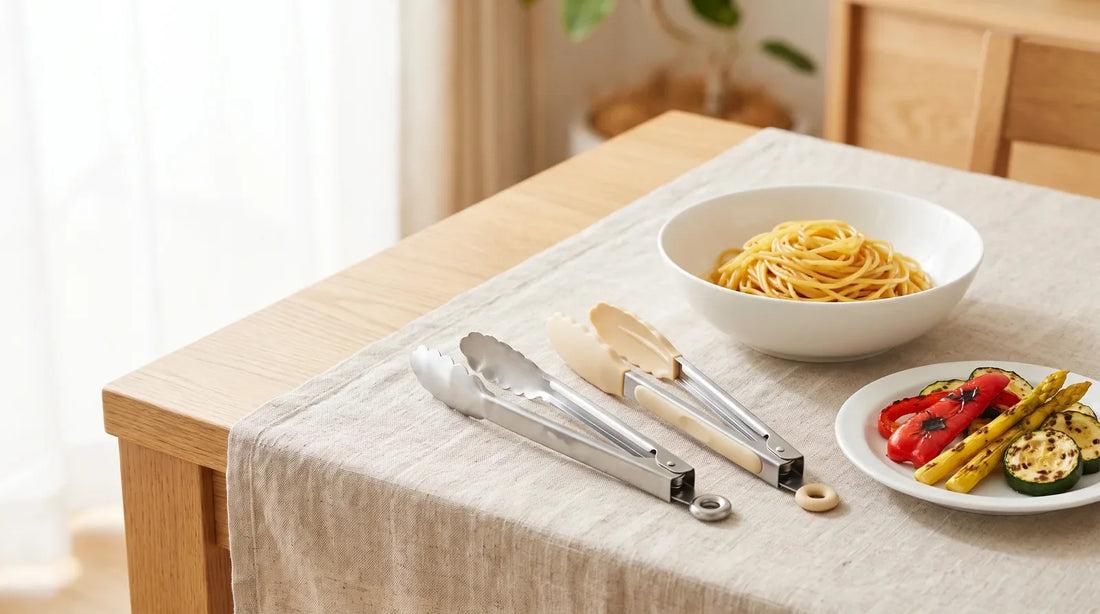 Stainless steel locking tongs and silicone-tipped tongs beside a ceramic bowl of pasta and a plate of grilled vegetables on a linen-covered light wood table