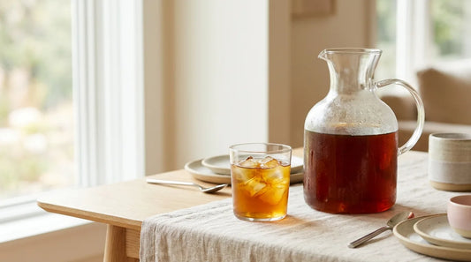 Large glass carafe of dark amber cold brew coffee beside an iced glass tumbler on a linen-covered light wood table