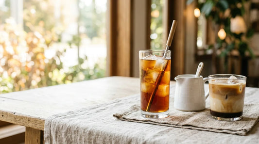 Tall glass of cold brew coffee over large clear ice beside a layered iced espresso drink on a light wood table with linen cloth