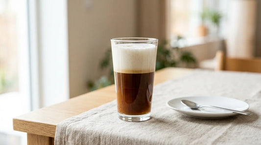 Tall clear glass latte showing clean espresso and microfoam milk layers on a light wood table with linen cloth
