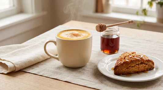 Maple latte in a cream stoneware mug beside a glazed scone and dark amber maple syrup jar on a linen-covered light wood table