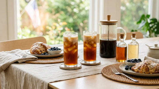 Two tall glasses of layered cold brew iced coffee beside a glass pitcher and syrup bottles on a linen-covered light wood table