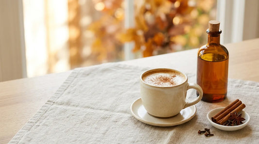Amber glass pumpkin spice syrup bottle beside a warm cream ceramic latte mug dusted with cinnamon, with whole cinnamon sticks and cloves on a linen-covered light wood table in autumn light