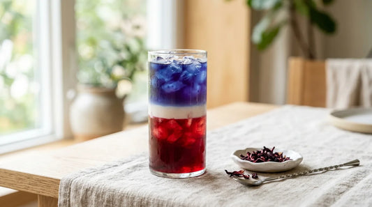 Tall clear glass with three distinct red, white, and indigo-blue layered iced coffee bands on a linen tablecloth with dried hibiscus flowers and a silver bar spoon