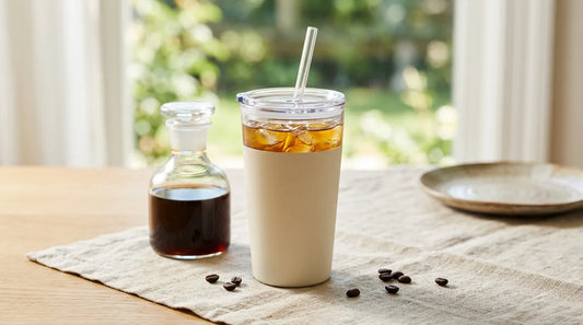 Reusable stainless steel iced coffee tumbler with straw lid filled with cold brew on a linen tablecloth beside a glass carafe