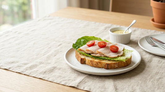 Open-face sourdough sandwich with romaine lettuce barrier and white ceramic sauce ramekin on a linen-covered light wood table, styled for a packed lunch
