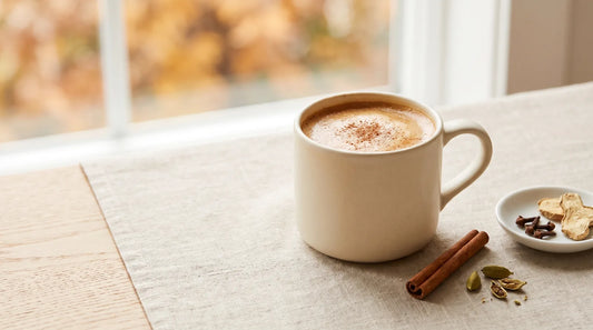 Spiced chai latte in a warm cream stoneware mug with cinnamon stick, cardamom pods, and cloves on a linen tablecloth, fall home café setting