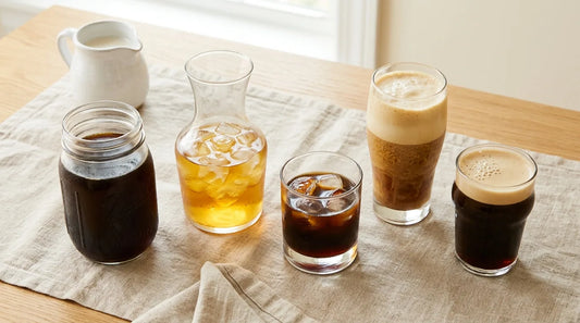 Five summer iced coffee drinks — cold brew, Japanese iced coffee, flash-chill espresso, shaken espresso, and nitro cold brew — arranged on a linen tablecloth on a light wood table