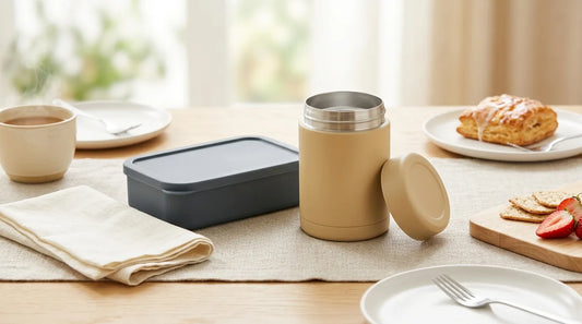Matte stainless-steel vacuum-insulated food jar and flat leakproof bento box on a linen tablecloth with a wooden serving board — café-style picnic lunch box setup